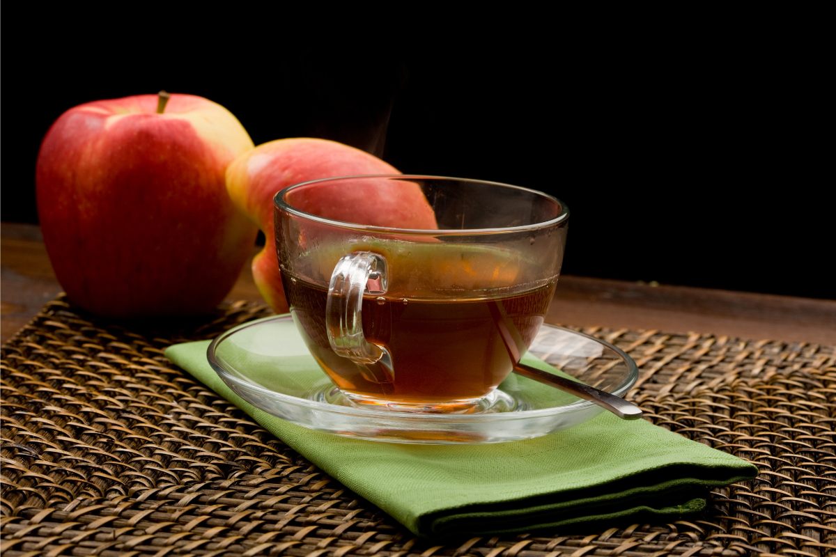 A cup of apple tea on a green napkin with two whole apples behind.