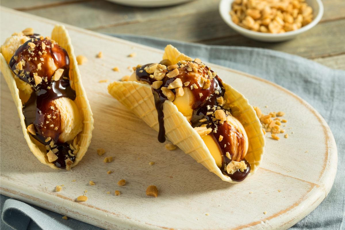 Two taco shaped biscuit shells filled with vanilla ice cream decorated with chocolate sauce and chopped peanuts.
