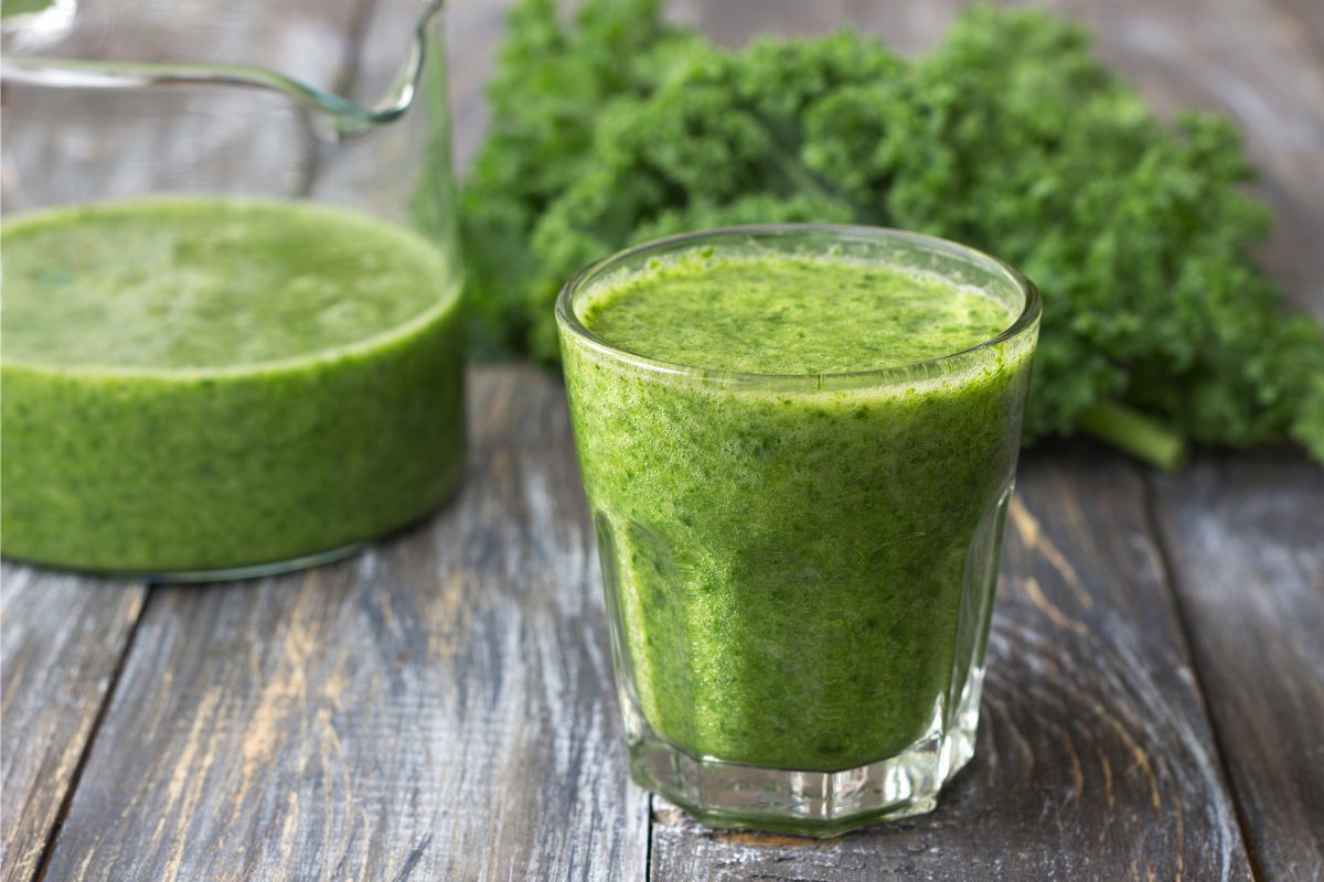 A glass filled with green smoothie on the foreground, a jar with some more smotthie and some kale on the background.
