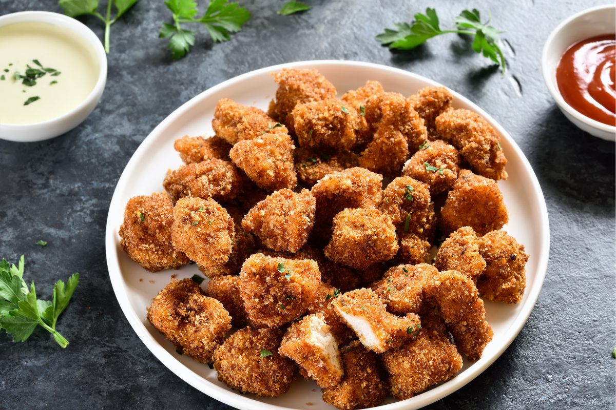 A plate of golden-brown breaded chicken bites garnished with herbs, served with white and red dipping sauces on the side.