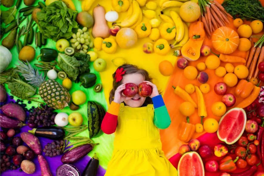 A girl covering her eyes with two apples and surrounded by a rainvow of fruit and vegetables.
