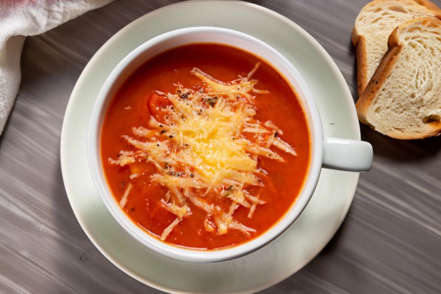 A cup with tomato and grilled cheese soup with some bread on the side.